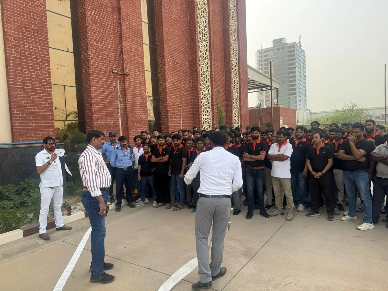 Fire safety instructor addressing a large group of factory staff through a megaphone during an on-site mock drill, a fire hose line running across the driveway