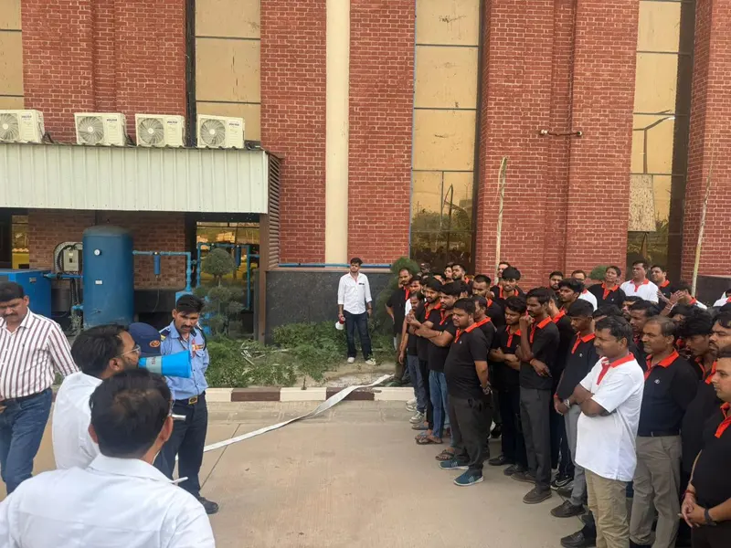 Employees in uniform lined up outside an industrial building beside a utility yard during a fire evacuation mock drill