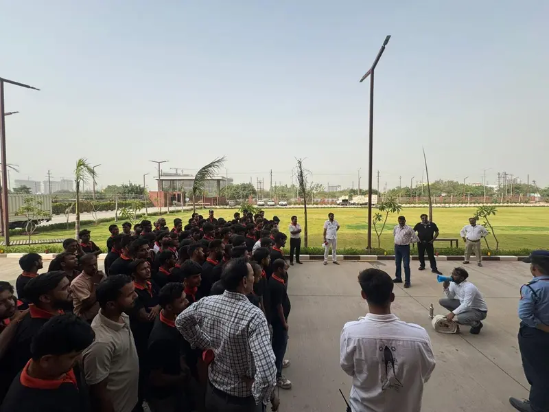 Wide view of factory employees mustered on a lawn-side assembly area during a fire mock drill, trainer kneeling beside a rolled fire hose