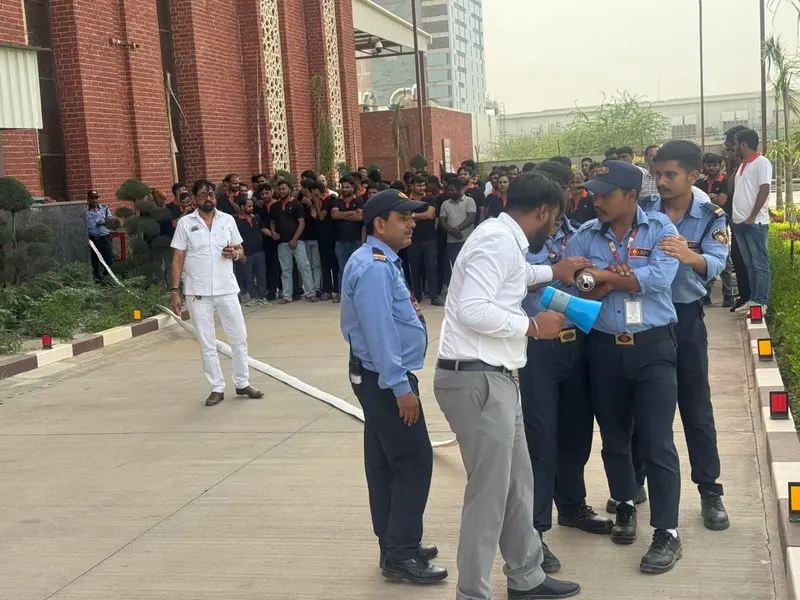 Security officers and factory employees gathered outside a building during a fire mock drill, with a fire hose laid out along the driveway