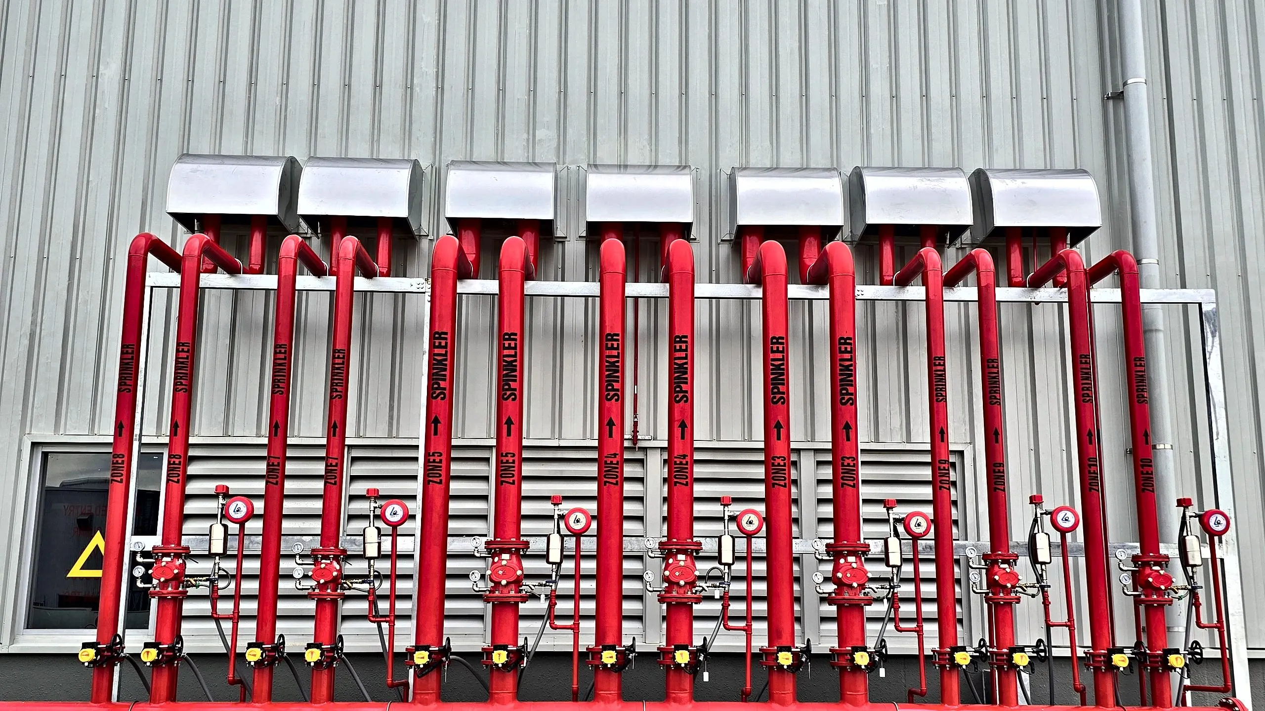 Row of red fire sprinkler and hydrant risers with control valves and gauges mounted along an industrial building wall