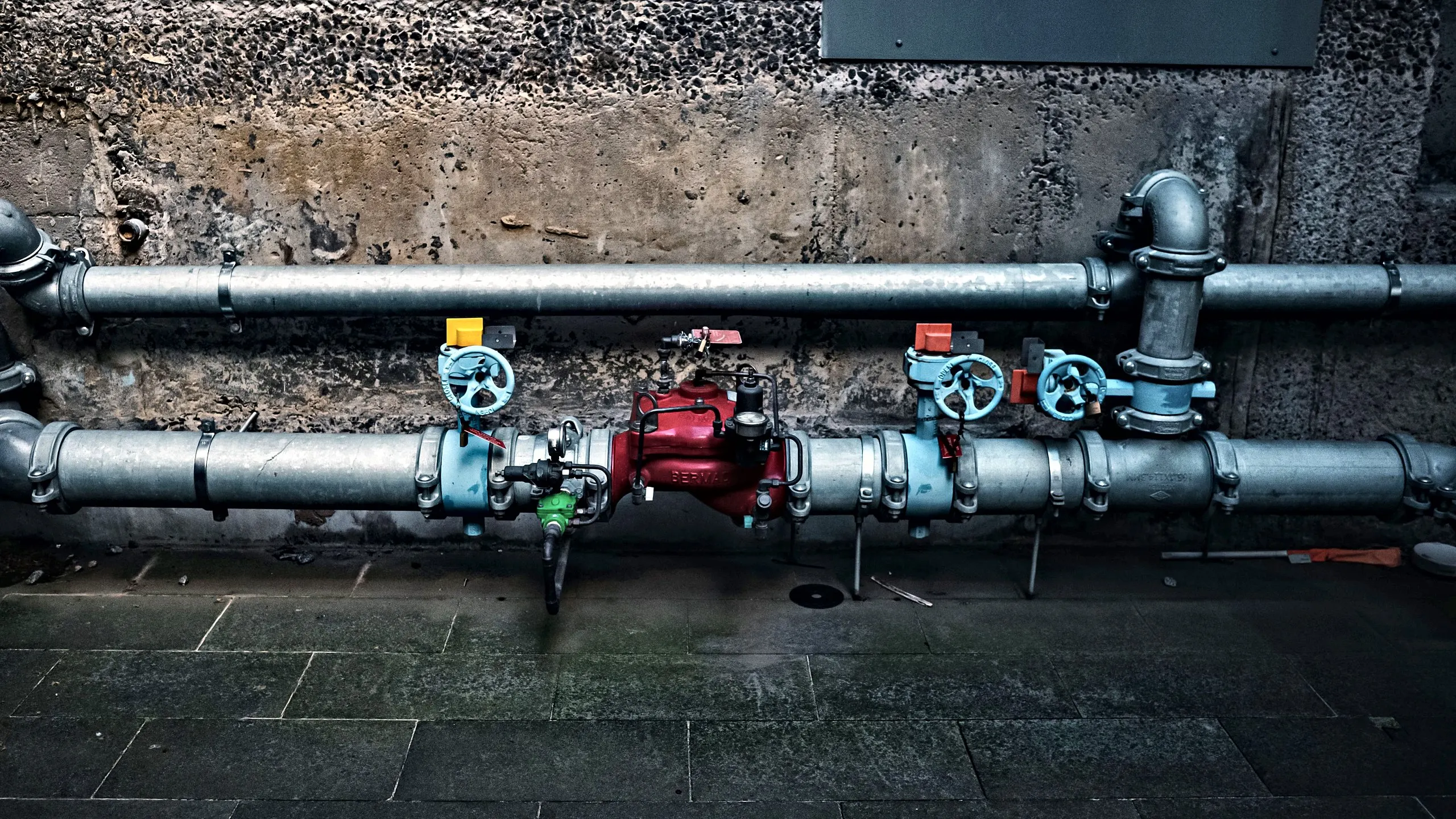 Fire water main with a red alarm check valve, isolation valves and pressure gauges along a concrete wall